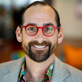 Derek Laney — smiling, red round glasses, grey blazer over a patterned shirt.